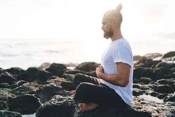 Foto op Plexiglas Concentrated male yogi dressed in comfortable wear meditating in lotus pose enjoying relaxation and concentration time at seashore rock, concept of holistic recovery and mental healthy lifestyle © BullRun