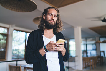Half length portrait of long haired Caucasian man with digital cellular technology looking at camera during free time, millennial male blogger 30s with modern mobile gadget posing indoors