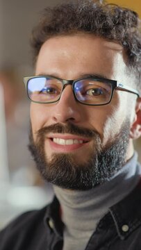 Vertical Portrait of a Happy Handsome Smart Male, Charmingly Smiling on Camera Indoors. Confident Successful Man Wears and Turtleneck and Glasses, Has a Bushy Beard and Short Curly Hair.