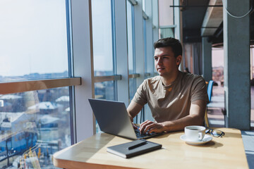 A young attractive male freelancer sits in the interior of a coffee shop and looks out the window....