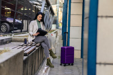 Full length portrait of a smiling successful business woman with a suitcase sitting and checking her documents in city.