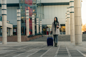 Full length portrait of a smiling successful business woman pulling a suitcase and carrying her documents and mobile phone.