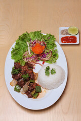 Vietnamese shaking beef with rice and vegetable are placed on a white plate, on a wooden table, served with chili sauce, salt pepper and lemon