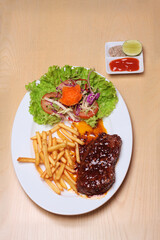 beefsteak with french fries and vegetable are placed on a white plate, on a wooden table, served with chili sauce, salt pepper and lemon