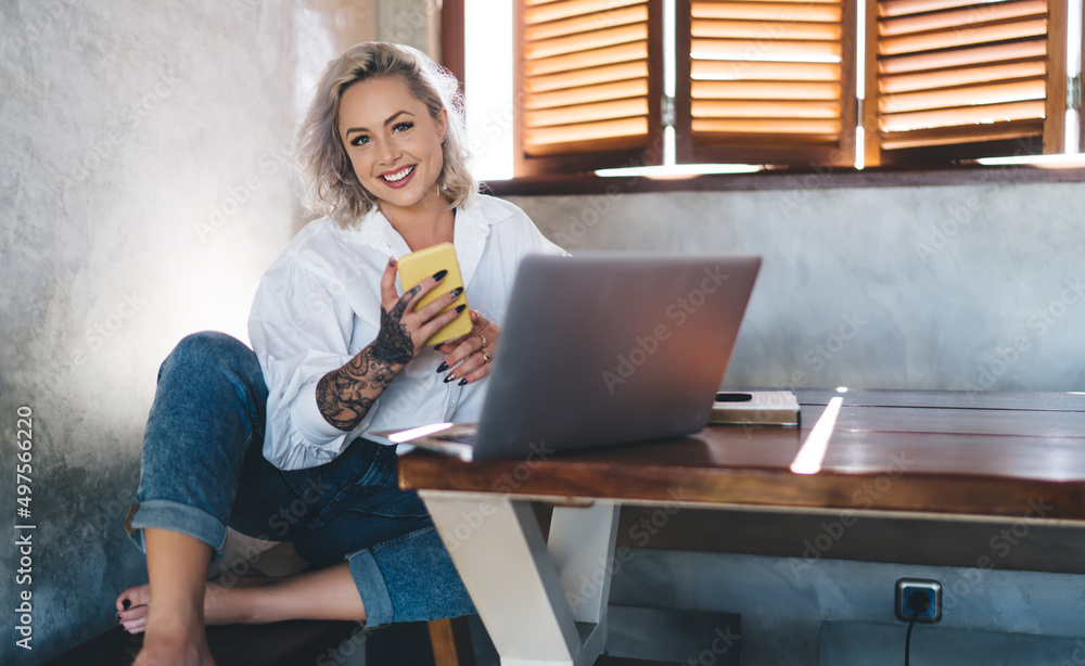 Poster portrait of funny female freelancer with mobile phone and netbook computer smiling at camera while d
