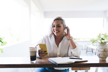 Portrait of happy female blogger with modern smartphone device and textbook smiling at camera enjoying time for creating content ideas, cheerful hipster girl with digital cellphone posing in cafe