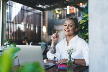 Half length portrait of cheerful Caucasian female digital nomad having break at table on cafe terrace, joyful woman freelancer sitting with laptop computer for doing distance job and smiling at camera