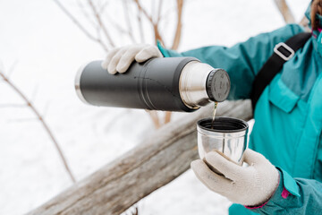 Thermos winter pour hot tea into a glass, hands holding a mug of hot drink, drink coffee in nature, hike winter mountain, breakfast in the forest, black thermos