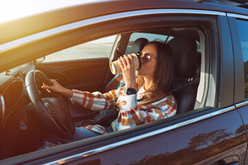 Young beautiful stylish woman driving her new car and drinking coffee on a sunny day. Concept for...
