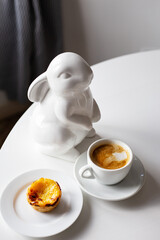 Easter Bunny stands near a cup of cappuccino and a cake on a plate, a white cup of coffee. Easter...