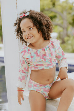 Portrait of smiling baby girl with afro hair style and sunglasses in a colourful swimsuit outfit posing by the pool. Afro-American and Afro-Brazilian children summer concept with copy space.