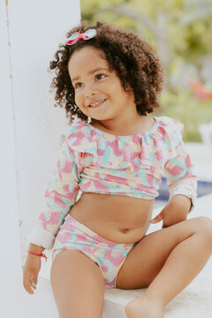 Portrait of smiling baby girl with afro hair style and sunglasses in a colourful swimsuit outfit posing by the pool. Afro-American and Afro-Brazilian children summer concept with copy space.