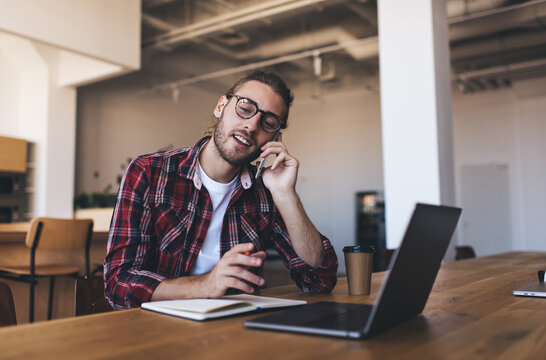 Millennial student with netbook computer and education textbook calling via cellular app for discussing homework while e learning, skilled programmer in spectacles have phone consultancy conversation