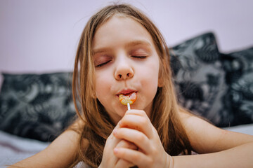 A beautiful, funny girl, a red-haired child, a sweet tooth, lies on the bed with a lollipop in her hands and wants to eat it.