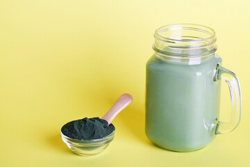 fresh green smoothie in glass bottle and spirulina powder with spoon on yellow background. useful habits, self care and healthy lifestyle. copy space