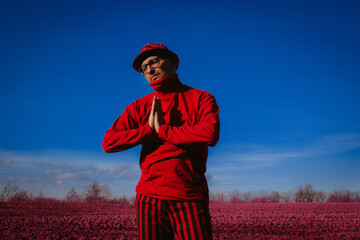 Fashionable man wearing red clothing, holding hands for prayer, posing outdoors with agricultural arable field and deep blue sky in the background