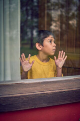lonely latin little boy leaning his hands behind a window pane, locked in a hut in the middle of...