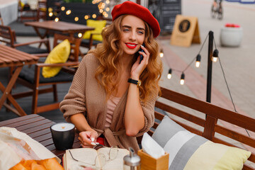 French clothing style, street fashion. A beautiful girl in a cardigan, red beret, and glasses walks through the old city and sits in a cafe. Spring mood and vibes