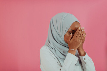 Modern African Muslim woman makes traditional prayer to God, keeps hands in praying gesture, wears traditional white clothes, has serious facial expression, isolated over plastic pink background