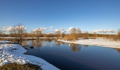 Fototapeta premium March sunny day by the river. A picturesque landscape, early spring, a river with snow-covered banks. The first thaws, the snow is melting.