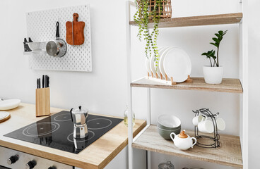 Modern shelving unit with tableware, pegboard and coffee maker on electric stove near white wall in kitchen
