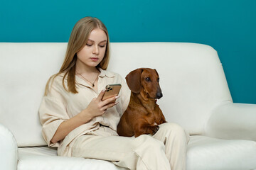 a pretty blonde girl is sitting on a white sofa, holding a red dachshund dog on her lap 