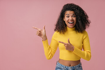 Excited African American woman wearing yellow sweater pointing finger on copy space isolated on pink background. Advertisement concept