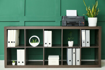 Shelf unit with modern printer, houseplant and folders near green wall in room