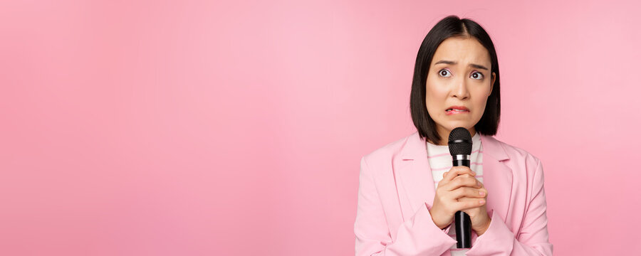 Anxious Asian Lady In Suit, Talking In Public, Giving Speech With Microphone On Conference, Looking Scared, Standing Over Pink Background