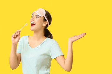 Pretty young woman with sleeping mask and toothbrush singing on yellow background