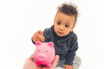 cute African American little boy sitting on the floor and putting a coin into the pink piggy bank with a serious face studio shot medium full shot white background copy space. High quality photo