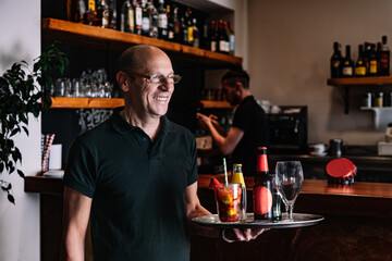 Mature, smiling waiter holding a tray with his hands. Worker in his small business carrying a tray of drinks.