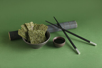 Bowl of tasty seaweed sheets, soy sauce and chopsticks on green background