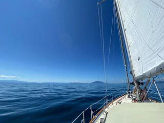 Wandaufkleber Blick auf ein Segelboot, das zusammen mit Segeln segelt und vom Wind gekippt wird. Blauer Himmel und Berge im Vordergrund © Sophia