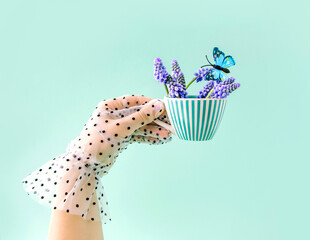 Spring creative arrangement of a woman's hand with a cup, flowers, and a butterfly on a blue background. Retro romantic summer aesthetic concept of the 80s and 90s. A surreal fashion idea for tea.