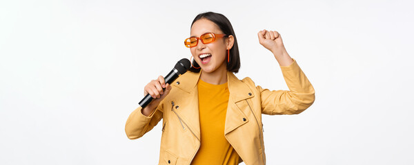 Stylish asian girl in sunglasses, singing songs with microphone, holding mic and dancing at...