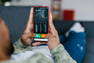 Close up view of young african american man lying on sofa using stock market app on smart phone.- Crypto concept with teenager boy selling currency coins