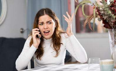 Wrathful young woman sitting at table in apartment and quarreling on phone.