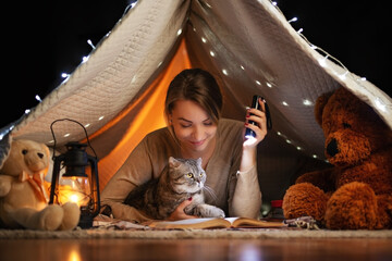 Obraz na płótnie Canvas Beautiful girl in a dark room with a cat in a tent reading a book with a lantern