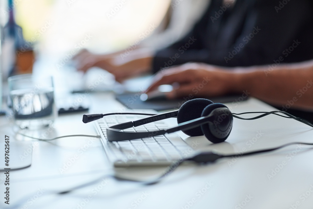 Aufkleber Get the assistance you need. Shot of a set of headsets lying on a desk with workers sitting in the background.