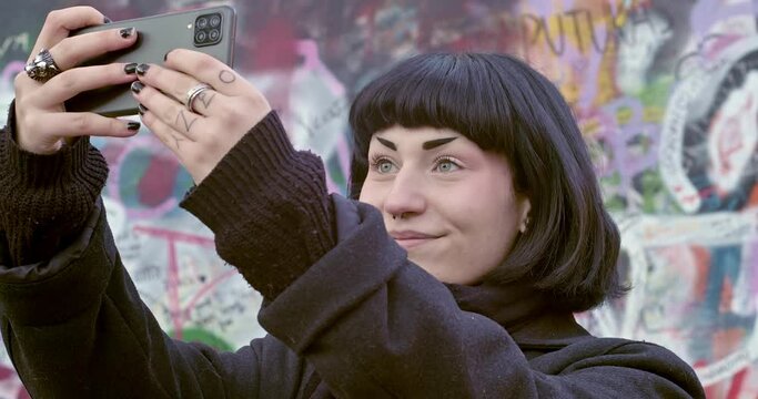 Interesting woman making mobile phone selfie in front of Lennon Wall full of graffiti in Prague