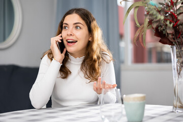Positive young woman sitting at table and having telephone conversation at home.