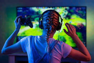 A gamer or a streamer girl at home in a dark room with a gamepad playing with friends on the networks in video games. A young man sits in front of a monitor or TV. - 497627449