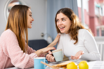 Two female friends have fun chatting while having tea at the table at home