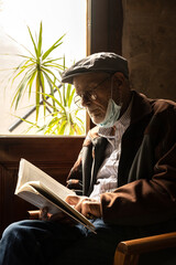 Elderly man in a cap and glasses reading a book by a door window.