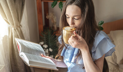 Young woman is reading a book and drinking coffee sitting by the window. Concept of morning enjoyment, leisure, pleasant pastime and relaxation