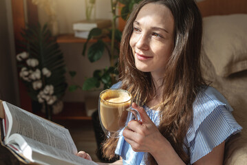 Young woman is reading a book and drinking coffee sitting by the window. Concept of morning enjoyment, leisure, pleasant pastime and relaxation