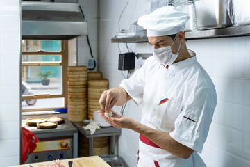 Male chef cook prepares dumplings and wearing face protective medical mask for protection from virus disease at restaurant kitchen.