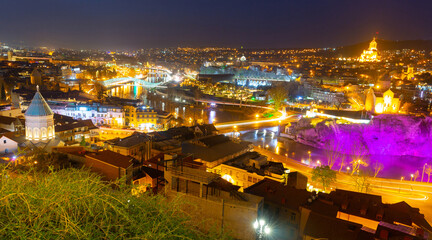 Fototapeta na wymiar Night spring cityscape of historical area of Tbilisi illuminated by colorful lights with view of modern bow-shaped bridge across Mtkvari River, Georgia