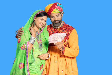 Happy indian couple wearing traditional colorful outfit holding indian rupees note against blue...